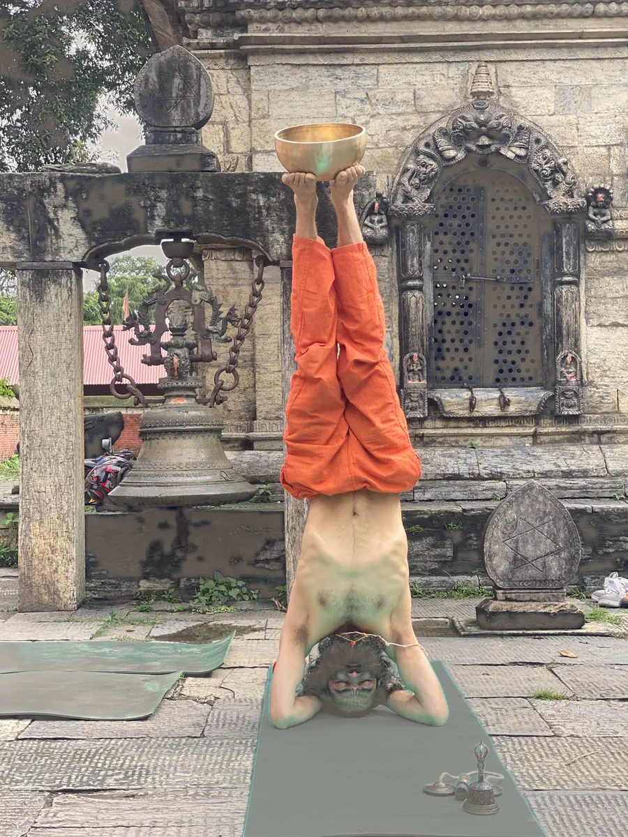 Yogi Ji balancing in a headstand while holding a bronze singing bowl aloft before a carved temple shrine.
