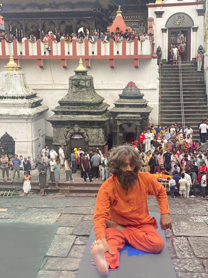Yogi Ji seated cross-legged in saffron dress before the steps of Pashupatinath, with pilgrims gathered behind.